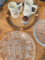 Group shot of two steins, stoneware jug, wicker tray, pressed-glass plate, partial view of textured glass plate