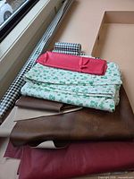 Stack of assorted fabrics showing gingham roll, green print cotton, brown vinyl, burgundy vinyl