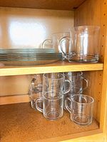 Cabinet shelves holding stacked clear plates, standard clear coffee mugs, and larger heavy glass mugs