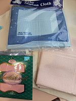 Group shot showing packaged baby afghan cloth, packaged pink Aida, folded pink piece, folded cream piece