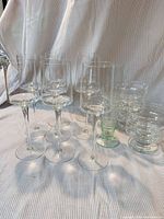 group of mixed clear glassware including tall stemmed wine glasses, stack of coupes, and green-tinted highball tumblers on fabric backdrop