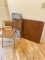 Two Samsonite folding chairs (one open, one folded) and square folding table top leaning against wall