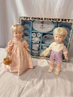 Two vinyl dolls standing in front of boxed china set