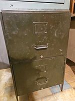 Front view of olive green two-drawer metal filing cabinet showing handles, label holders, and overall wear