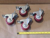 Four red 3-inch swivel caster wheels with brakes laid out, tape measure shows scale
