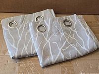 Two folded ash grey curtain panels with white branch pattern and metal grommets displayed on cardboard surface