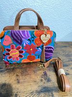 Front of handbag showing embroidered panel, top handles, heart tassel charm, detachable strap