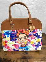 Front view showing Frida illustration, multicolor canvas, tan faux-leather top section, wooden heart charm and tag attached