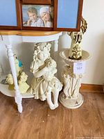 Overall view of statues beneath table: girl with lamb, pedestal, angel, Virgin Mary, child figures