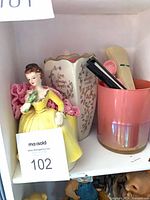 Shelf view showing lady figurine, vase, pink tumbler with fans and crocheted piece