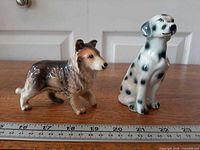 Collie and Dalmatian figurines side by side on table with ruler for scale