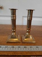 frontal view of both brass candlesticks showing square bases and cylindrical stems next to ruler