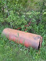 Full view of rusted steel roller lying in grass with visible crack at end