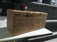 Front and top of sealed APC Back-UPS box showing branding, product illustration, and intact tape