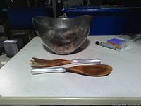 Metal salad bowl with two serving utensils on table