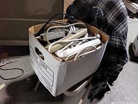 Cardboard file box on chair filled with assorted power strips, cords visible.