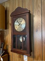 Front view of oak wall clock on paneled wall, dial and pendulum window visible