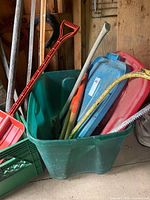 Green tote containing assorted tools including red snow shovel, red rake head, axe handle and bow saw