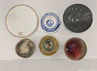 Top view showing six of the pieces: bone china plate, blue pattern cup on saucer, black bowl, sand-tone plate, signed bowl, red-interior vessel