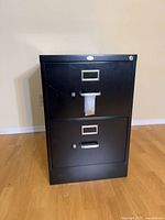 Front view of black 2-drawer filing cabinet showing handles, label holders, and lock