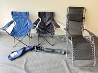 Two open folding camp chairs, one blue, one black, collapsed chair bags, and zero-gravity lounger
