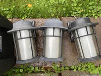 Three matching cylindrical outdoor wall light fixtures on paving stones, showing overall condition
