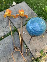 Blue glass gazing ball on stand and two orange glass flower stakes laid on stone