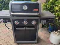 Front view of Coleman Revolution 3-burner propane grill showing controls, hood, thermometer, side shelves, and propane tank