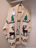 Front view of cream cardigan with brown moose and green tree motifs hanging on hanger