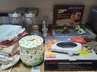 Overview of lot showing boxed appliances, ceramic jar, glass canisters, grinders, linens, cords