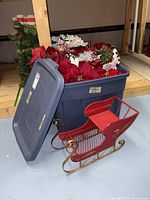 Blue Rubbermaid tote filled with artificial poinsettias; red metal sleigh displayed in front