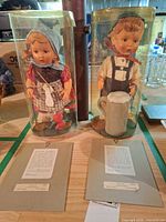 Front view of both Hummel Village series dolls in clear display tubes with certificates and accessories visible