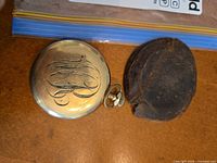 Back of monogrammed gold tone pocket watch beside leather pouch