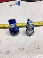 Front view of both blue and white porcelain Dutch boy and girl shakers with measuring tape showing height