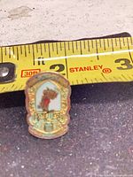 Front of small gold tone lapel pin with red and white enamel against measuring tape showing under 1 inch