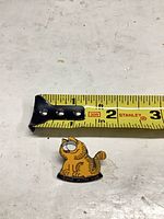 Front of enamel cartoon cat pin beside measuring tape showing about 1 inch width