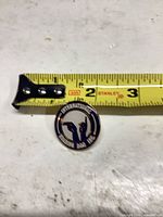 Front view of enamel pin beside tape for scale