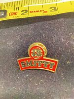 Front view of gold-tone red enamel Smitty 18 lapel pin next to ruler