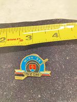 Front view of enamel lapel pin next to measuring tape showing approx 1 in width