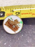 Front of circular enamel pin beside tape measure showing under 1-inch diameter