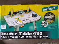 Box with image and text showing Wolfcraft Router Table 490, illustrating the assembled router table with adjustable fences and handles.