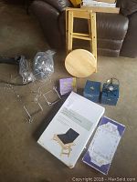 Overall lot arranged on floor showing laptop tray in box, disassembled bench parts, candle holders, meditation calendar, letter set, decorative stand and blue box with clock.
