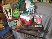 Photo of garden supplies lot showing green coiled hose with nozzle, red cooler, trimmers, and various accessories on table with gray cover behind