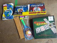 Photo showing all items in the lot: Cross Cribb strategy game box, two Magnetix toy sets, wooden cribbage board, children's toy racket and ball, and Trivial Pursuit electronic game.