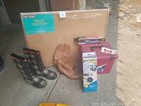 Lot items arranged outdoors showing plaid rain boots, brown Boulet western boots, Staples cork bulletin board in cardboard sleeve, and other small items.