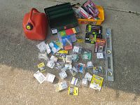 Overview photo of the entire lot showing green and yellow toolboxes, red gas container, assorted small packaged items including drill bits, adhesives, repair kits, screws and hooks scattered on the ground.