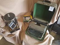 Remington typewriter in green case next to travel iron, small radio, and other small vintage items on beige fabric.