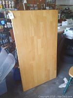 Wooden rectangular tabletop, light natural wood finish, standing vertically in a storage area.