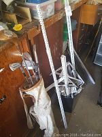 Golf clubs in white golf bag alongside metal shelving parts and poles leaning against wall.