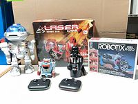 Overall shot showing all five items: boxed Khet 2.0 game, Robotix kit, silver robot, small RC robot, Darth Vader figure, two controllers